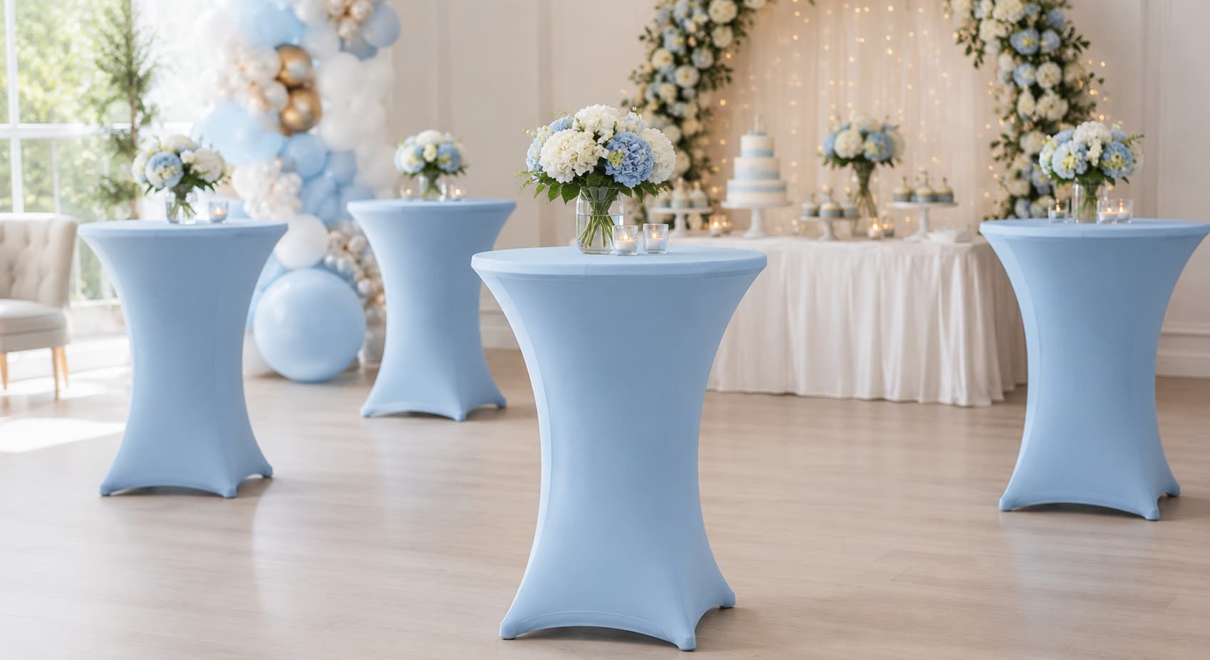 Vier statafels met babyblauwe statafelrokken in een feestzaal, met bloemboeketten, ballonnenboog en bruidstaart op de achtergrond